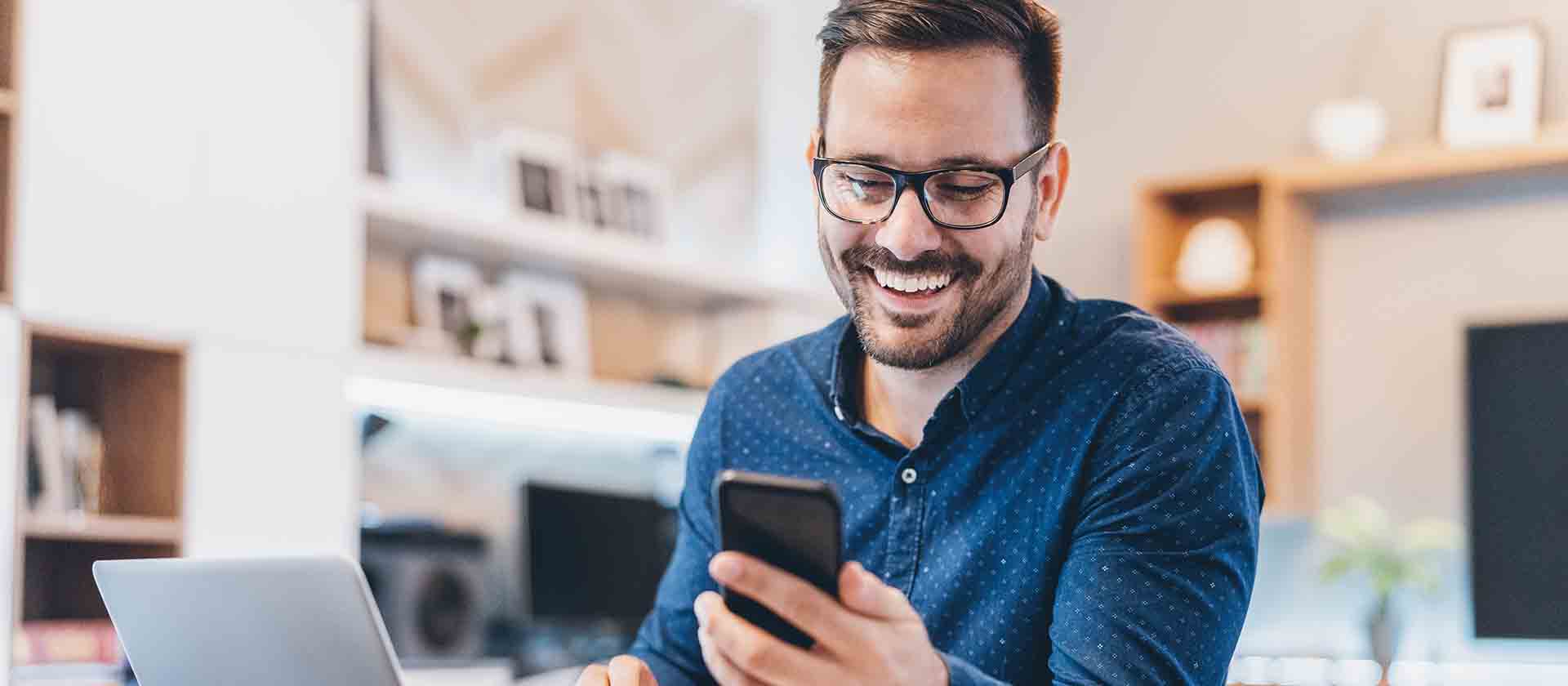 Ein lächelnder Mann mit Brille nutzt sein Handy und strahlt Freude aus.